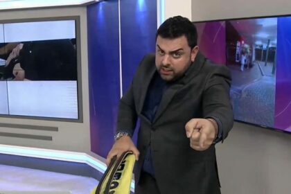 Macedão segura um chinelo gigante amarelo e aponta para o telespectador no cenário do Tá na Hora no SBT