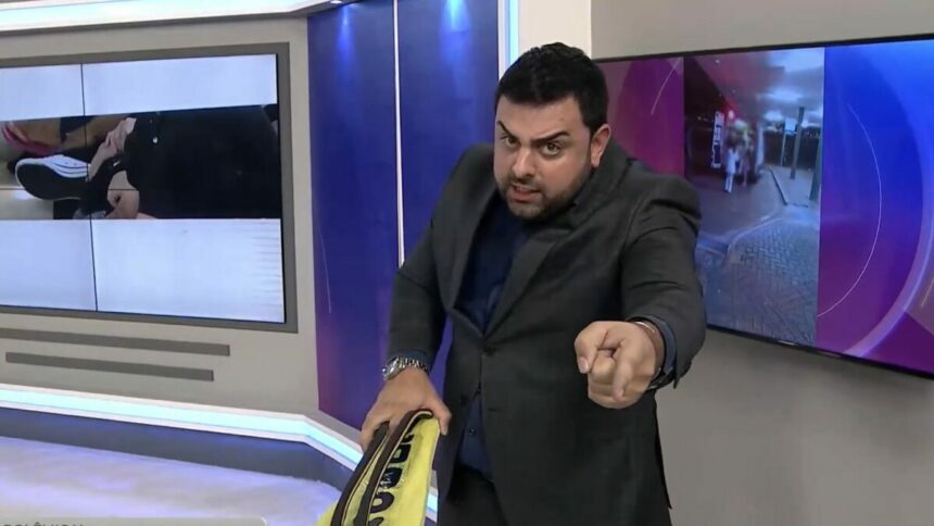 Macedão segura um chinelo gigante amarelo e aponta para o telespectador no cenário do Tá na Hora no SBT