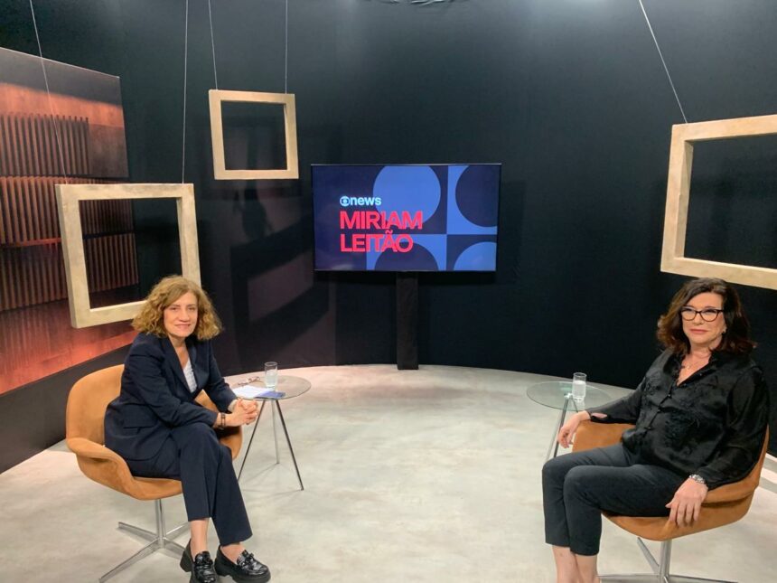 Miriam Leitão sentada em uma cadeira no cenário da Globo News ao lado da presidente da Petrobras, Magda Chambriard