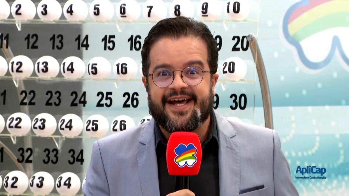Apresentador do Pernambuco da Sorte em frente ao painel com as bolas do sorteio de 11 de maio. Ele segura um microfone vermelho