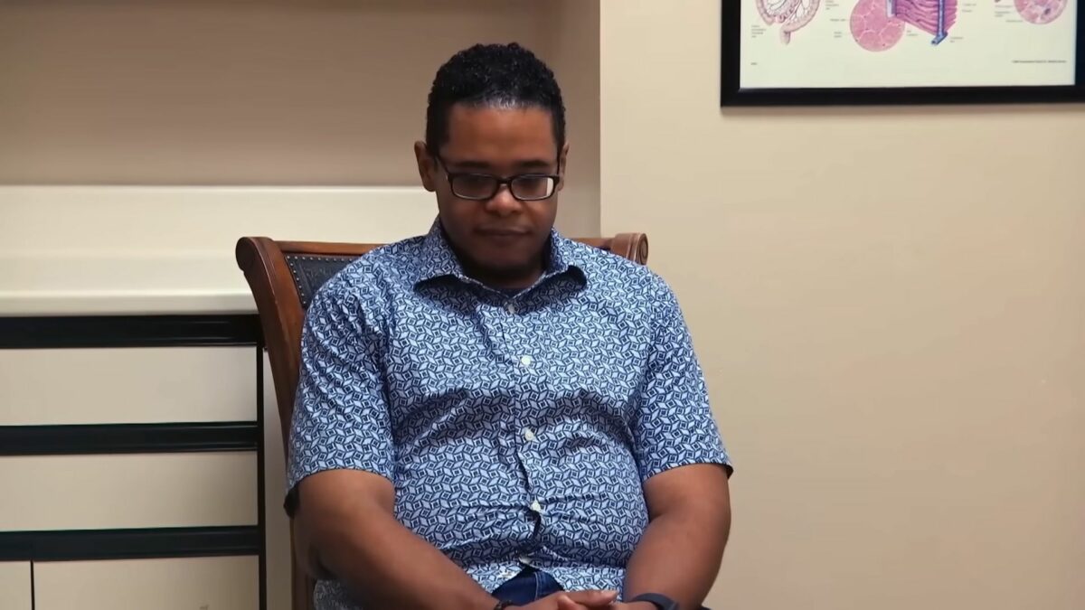Brandon sentado em uma cadeira e olhando para o chão. Ele veste camisa de botão e está bem mais magro
