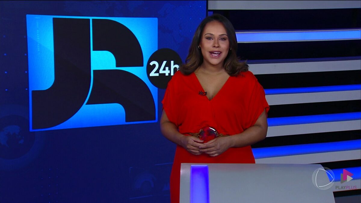 Salcy Lima de vestido vermelho apresentando o Jornal da Record. Ela está em frente ao telão vertical com o logo do JR 24h