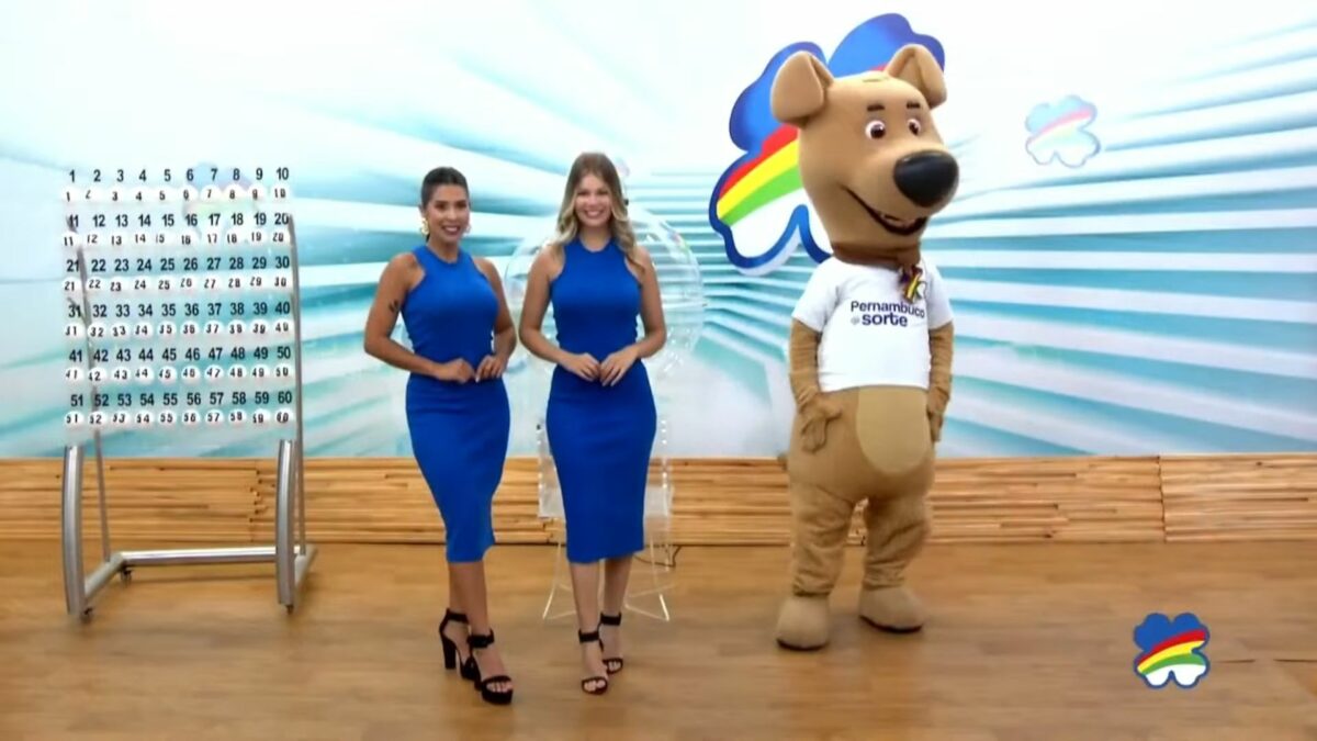 Assistentes de palco vestem azul, posicionadas ao lado do painel com as bolas e o mascote do sorteio do Pernambuco dá Sorte