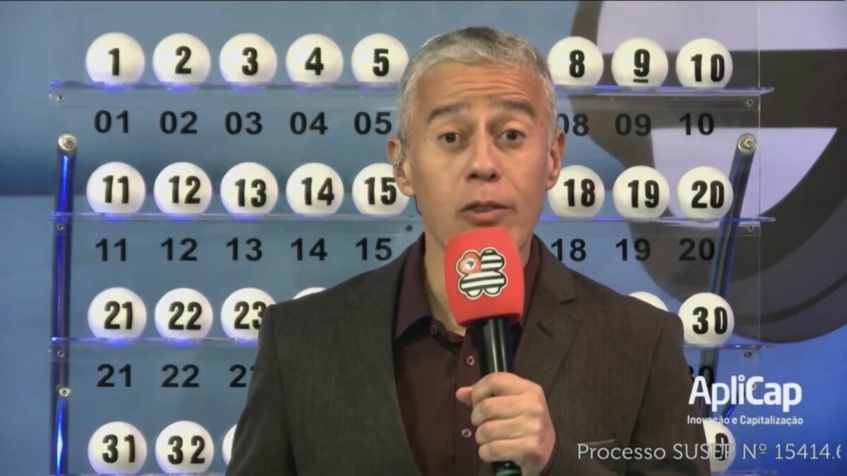 Apresentador do São Paulo da Sorte em frente ao painel com as bolas do sorteio de 11 de maio. Ele segura um microfone vermelho