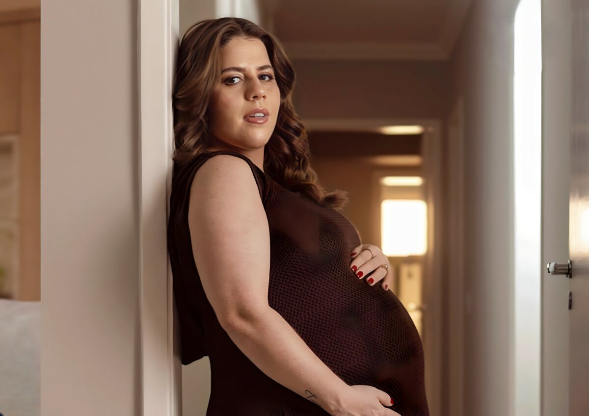 Tati Machado com a mão na barriga ainda grávida. Ela veste um vestido marrom e está encostada em uma parede