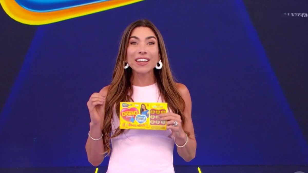 Patricia Abravanel sorrindo e segurando o bilhete do quarto sorteio da Tele Sena de Mães 2025. Ela veste um vestido branco.