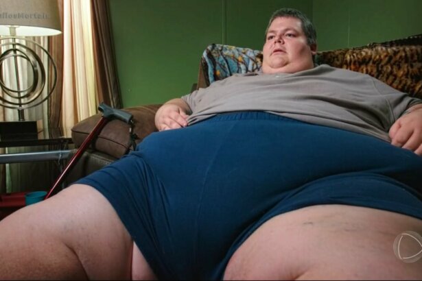 Aaron extremamente obeso, pesando 326 quilos, sentado em um sofá. Suas pernas e barriga se confundem
