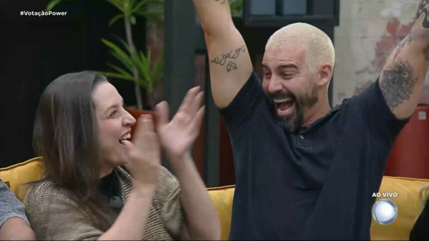 Carol e Radamés celebram vitória na Prova dos casais. Radamés levanta os braços, sentado no sofá, enquanto Carol bate palmas