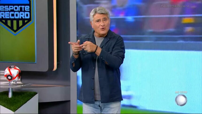 Cleber Machado sorrindo no centro do cenário de transmissão de jogo na RECORD; Narrador está em pé!