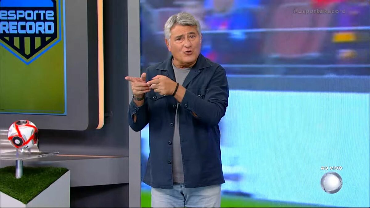 Cleber Machado sorrindo no centro do cenário de transmissão de jogo na RECORD; Narrador está em pé!