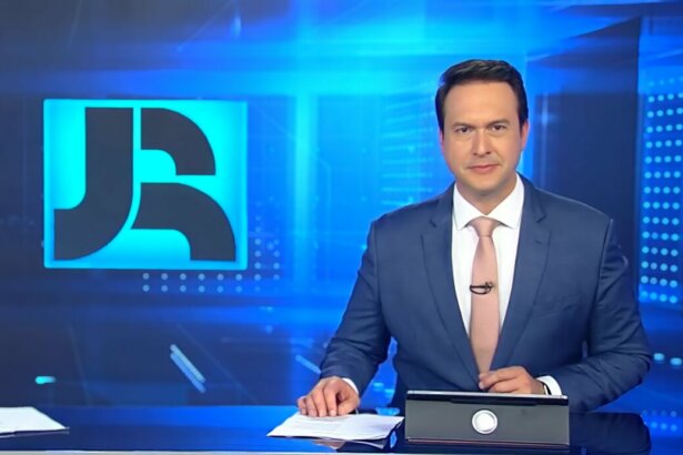 Eduardo Ribeiro sentado na bancada do Jornal da Record vestindo terno e gravata Salmon. Ao fundo o telão do JR com o logo do telejornal