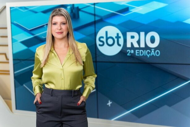 Livia Mendonça em pé, de blusa verde e calça cinza de alfaiataria no cenário do SBT Rio 2ª Edição