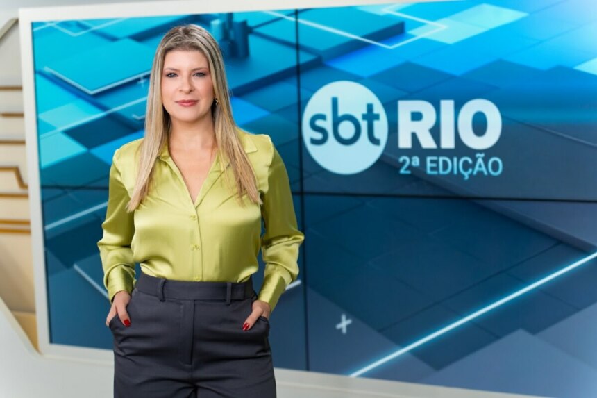 Livia Mendonça em pé, de blusa verde e calça cinza de alfaiataria no cenário do SBT Rio 2ª Edição