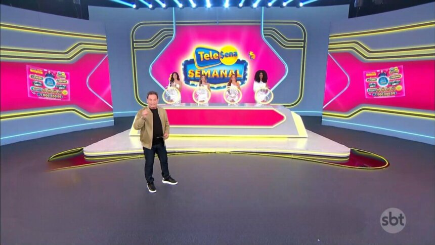 Luis Ricardo caminha no cenário do sorteio da edição 55 da Tele Sena Semanal. O cenário está com artes gráficas rosa, cor do bilhete