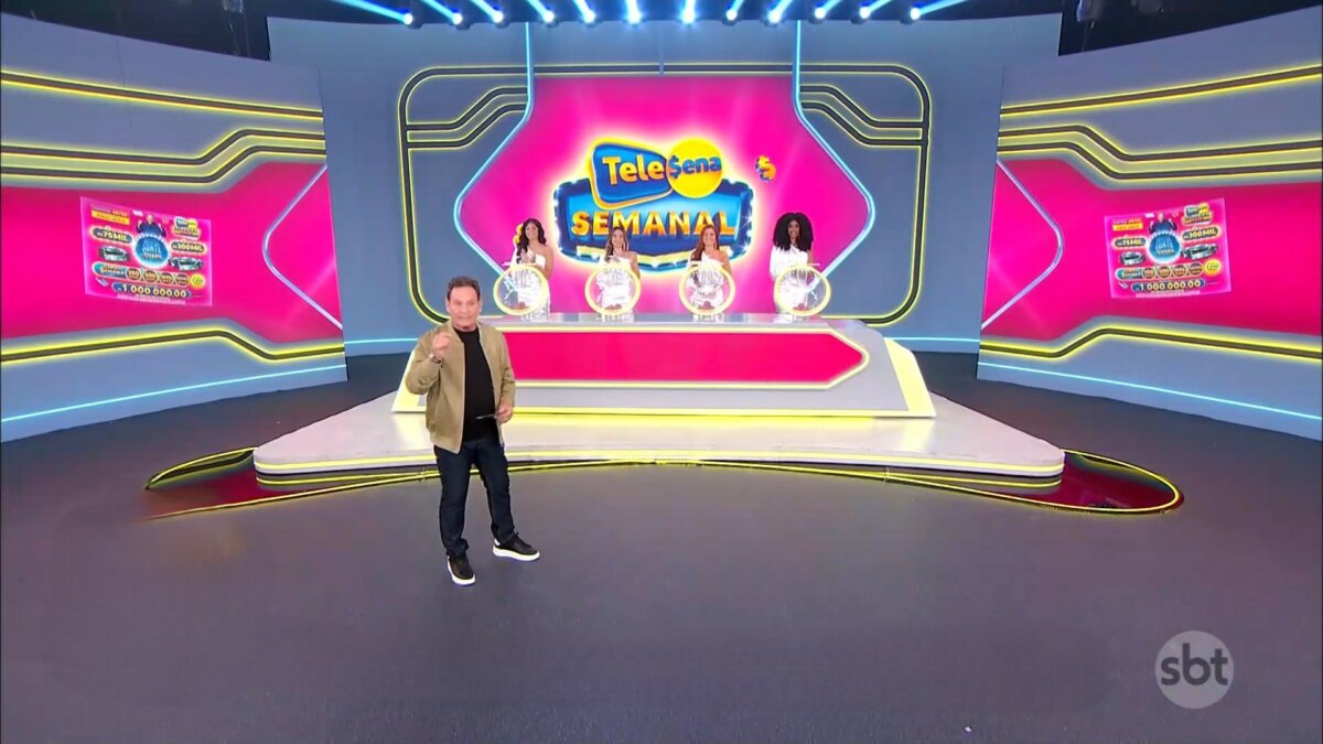 Luis Ricardo caminha no cenário do sorteio da edição 55 da Tele Sena Semanal. O cenário está com artes gráficas rosa, cor do bilhete