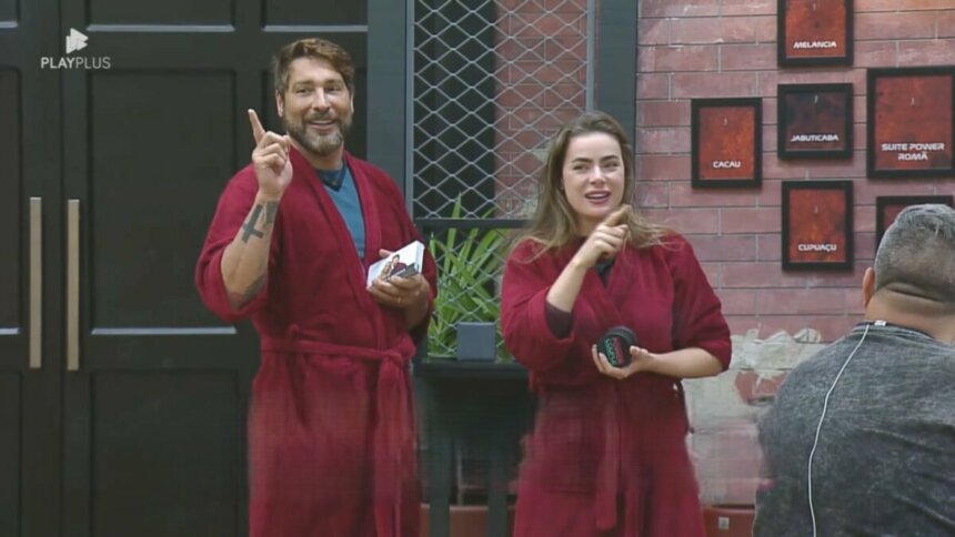 Victor e Rayanne seguram as placas dos quartos do Power Couple Brasil 7. Eles vestem roupão vermelho do casal Power e estão sorrindo