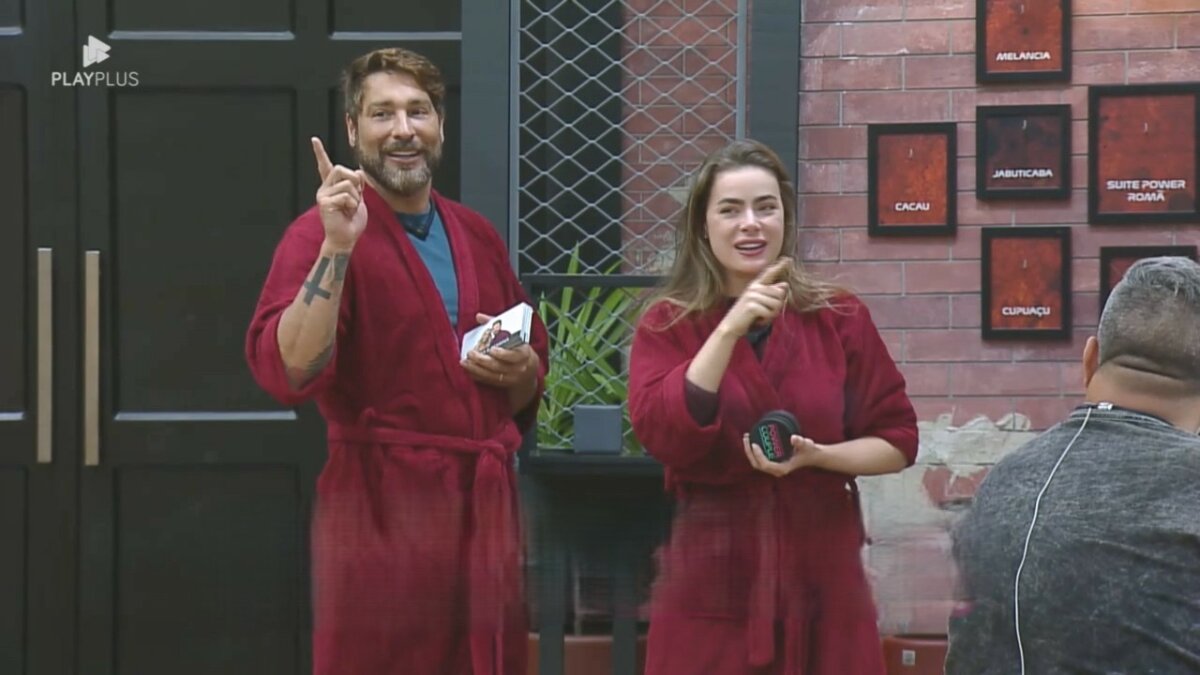 Victor e Rayanne seguram as placas dos quartos do Power Couple Brasil 7. Eles vestem roupão vermelho do casal Power e estão sorrindo