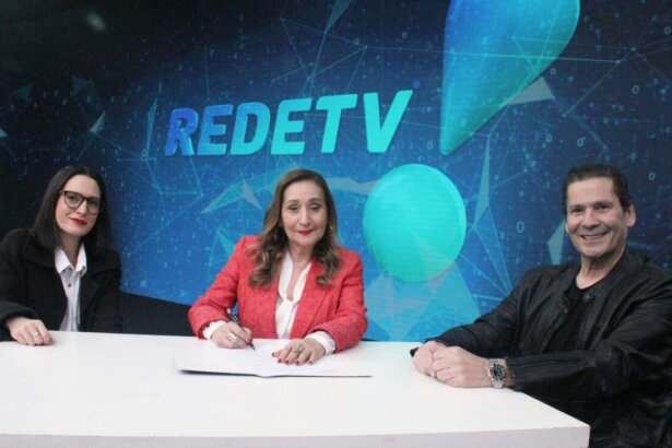 Sônia Abrão no momento em que assina renovação de contrato com a RedeTV! até 2029 com Andrea Dallevo (à esq.) e Elias Abrão (à dir.) por trás dos três o supertelão de LED