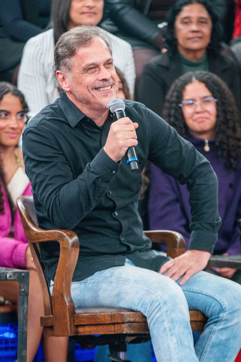 Ator Guilhermes Fontes sentado na cadeira de madeira do Altas Horas segurando o microfone da Globo