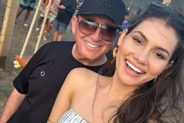 Amado Batista de óculos escuro atrás da esposa Calita Franciele que sorrir. Eles estão em uma rua