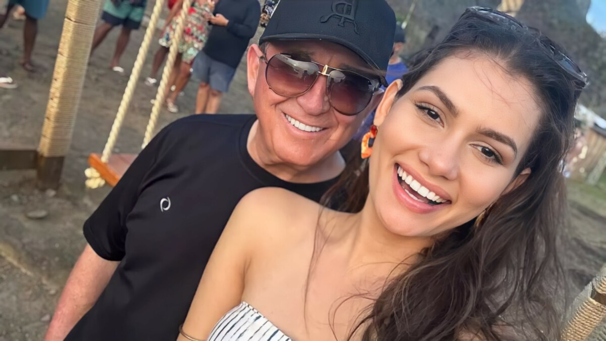 Amado Batista de óculos escuro atrás da esposa Calita Franciele que sorrir. Eles estão em uma rua