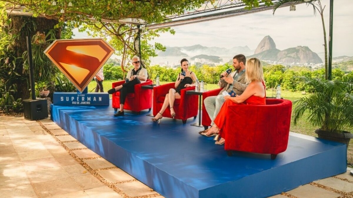 David Corenswet, Rachel Brosnahan e James Gunn sendo entrevistados com o logo do filme Superman ao lado. Ao fundo a paisagem do Rio de Janeiro