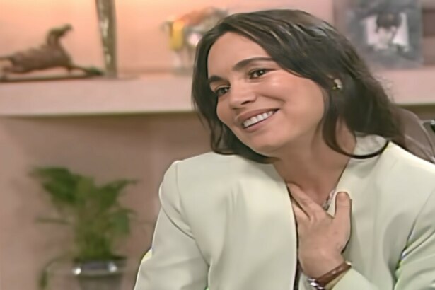 Helena, Regina Duarte, com a mão no peito em ar de felicidade. Ela está sentada no escritório de Carlos em cena de História de Amor