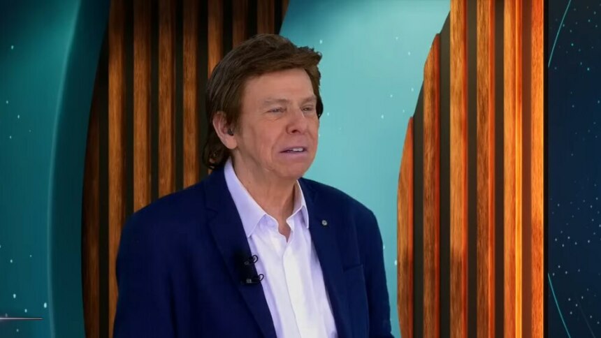 Nelson Rubens de terno e camisa branca no cenário do TV Fama olhando sério para o lado direito