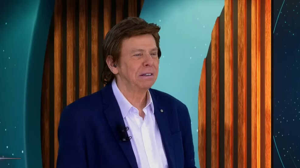 Nelson Rubens de terno e camisa branca no cenário do TV Fama olhando sério para o lado direito