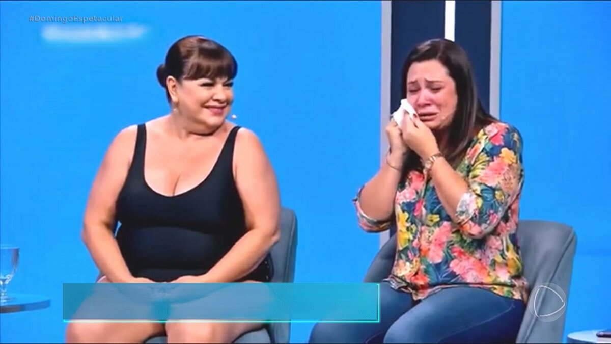 Mulher criada por IA, usa maiô e olha para o lado onde uma entrevistada chora no Programa Marisa Maiô