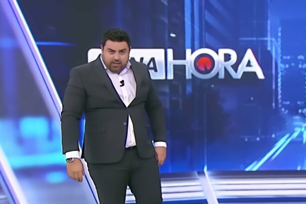Macedão olha por baixo no cenário do Tá na Hora. Ele veste terno preto e apertado com botão quase saindo