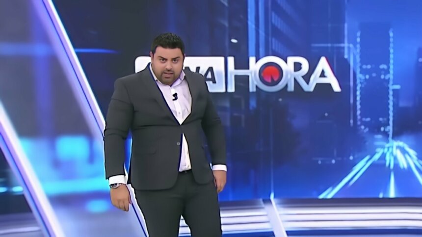Macedão olha por baixo no cenário do Tá na Hora. Ele veste terno preto e apertado com botão quase saindo