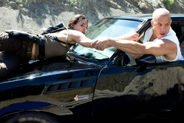 Toretto segura na mão de sua parceira que está pendurada no capô de um carro em cena de Velozes e Furiosos 4