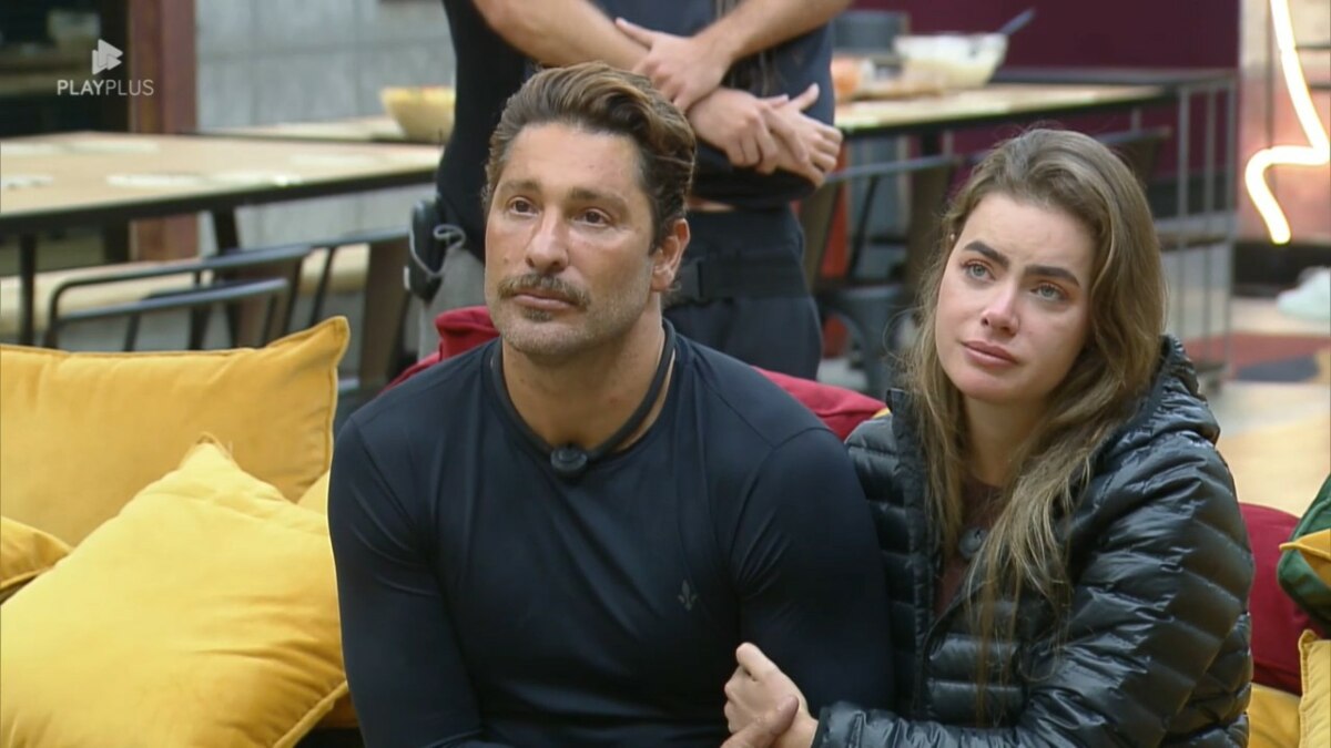 Victor e Rayanne sentados no sofá amarelo da Mansão olham atentamente para o telão onde está Felipe Andreoli antes de anunciarem veto na Prova dos Casais