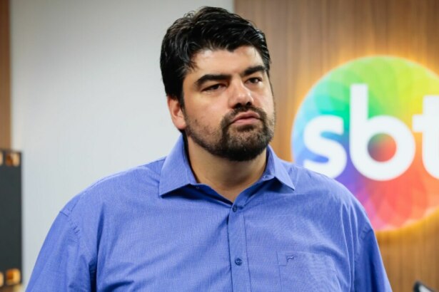 André Azeredo vestindo uma blusa social azul, sorridente, em foto posada em frente ao logo do SBT