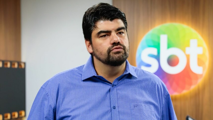 André Azeredo vestindo uma blusa social azul, sorridente, em foto posada em frente ao logo do SBT