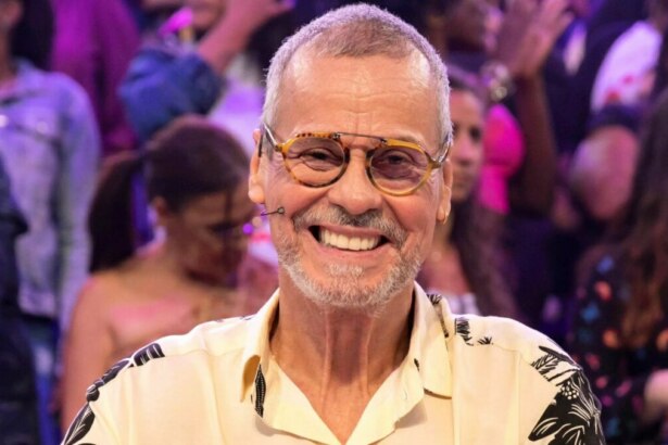 Carlinhos de Jesus sorridente e sentado na bancada de júri da Dança dos Famosos