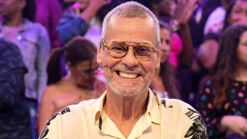 Carlinhos de Jesus sorridente e sentado na bancada de júri da Dança dos Famosos