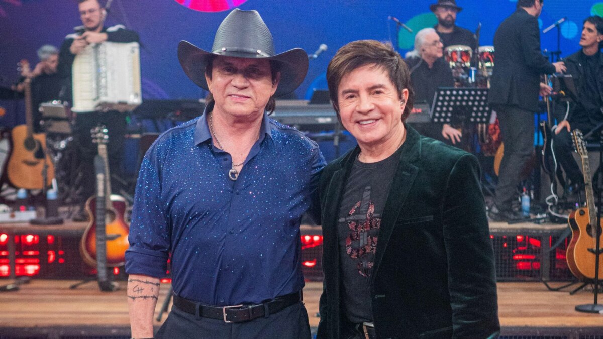 Cantores Chitãozinho e Xororó abraçados, em foto posada, em frente a banda, no cenário do Altas Horas