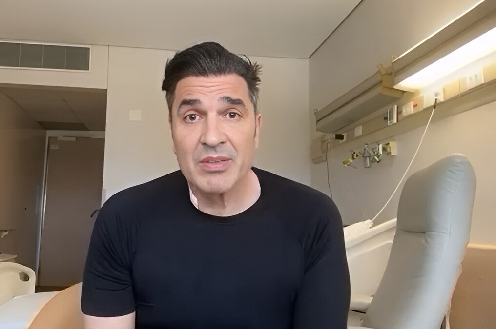 Edu Guedes veste camisa preta e está sentado em uma maca de hospital
