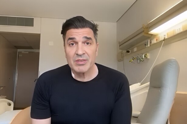 Edu Guedes veste camisa preta e está sentado em uma maca de hospital