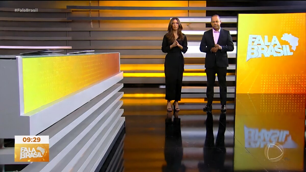 Paloma Poeta e Luiz Fara Monteiro em pé, ao lado do telão do Fala Brasil