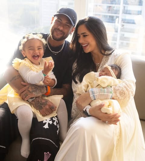 Neymar, Mavie, Bruna e Mel. O jogador veste preto, mãe e filhas vestem amarelo e todos estão sorridentes, sentados em um sofá. Bruna segura Mel nos braços