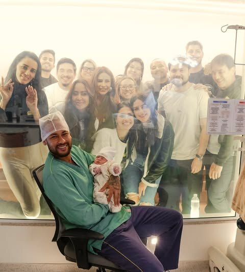 Neymar segura Mel nos braços e mostra aos familiares que estão fora do quarto vendo por uma janela de vidro