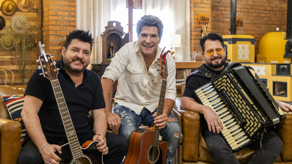Bruno e Marrone sentados no sofá segurando guitarra e sanfona. Por trás deles o apresentador Daniel nos bastidores do Viver Sertanejo