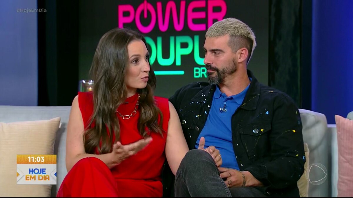 Carol e Radamés sentados no sofá do Hoje em Dia. Carol veste um vestido vermelho e Radamés de jaqueta preta e camisa azul