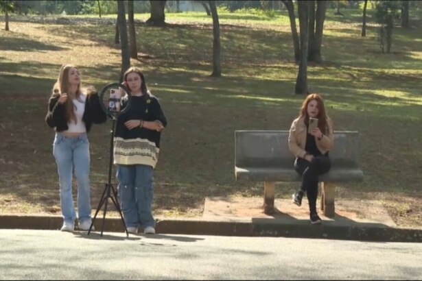 Meninas gravam vídeo em um parque, com um tripé apoiando um celular enquanto outra menina sentada em um banco mexe em seu smartphone em cena do quadro E se fosse Você? do Hoje em Dia