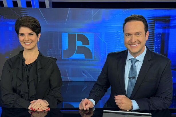 Mariana Godoy e Eduardo Ribeiro sorridentes, sentados na bancada do Jornal da Record, eles vestem roupas pretas