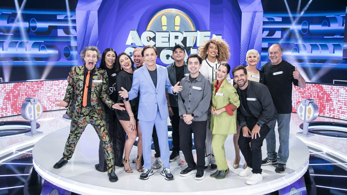 Tom Cavalcante reunido com o elenco do Acerte ou Caia! no centro do palco do programa todo iluminado. Eles vibram e estão sorridentes
