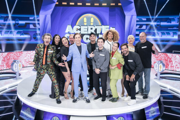 Tom Cavalcante reunido com o elenco do Acerte ou Caia! no centro do palco do programa todo iluminado. Eles vibram e estão sorridentes